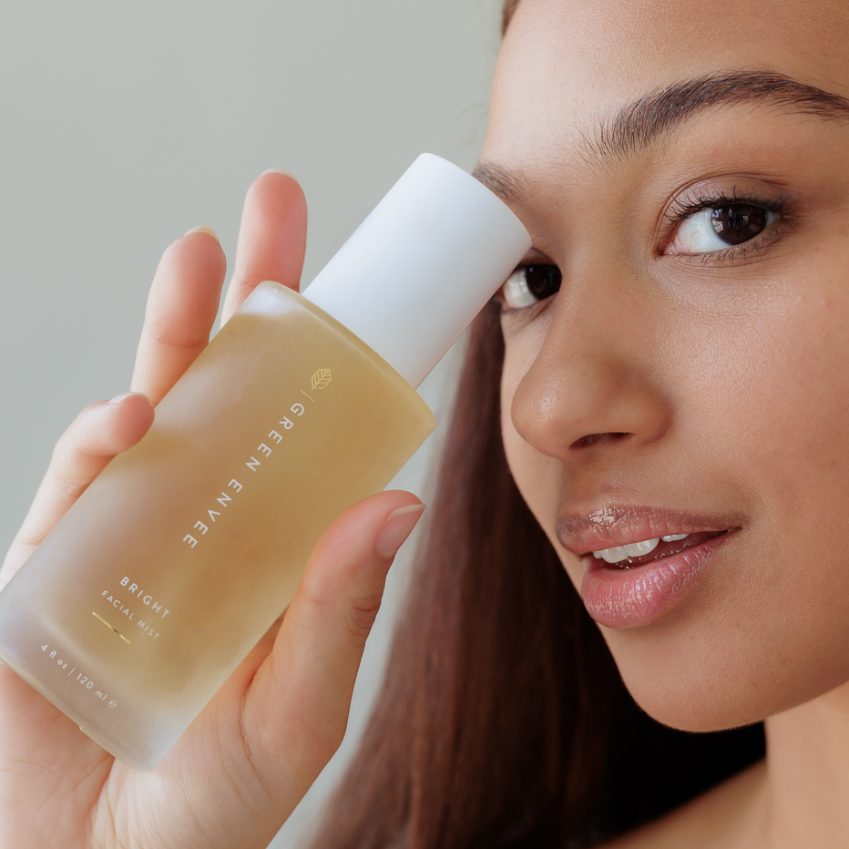 Woman holding a bottle of 'Green Envee' bright mist skincare product near her face.
