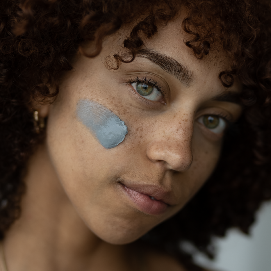 Close-up of a woman with a charcoal cosmetic cream product on her face against a blurred background.  
