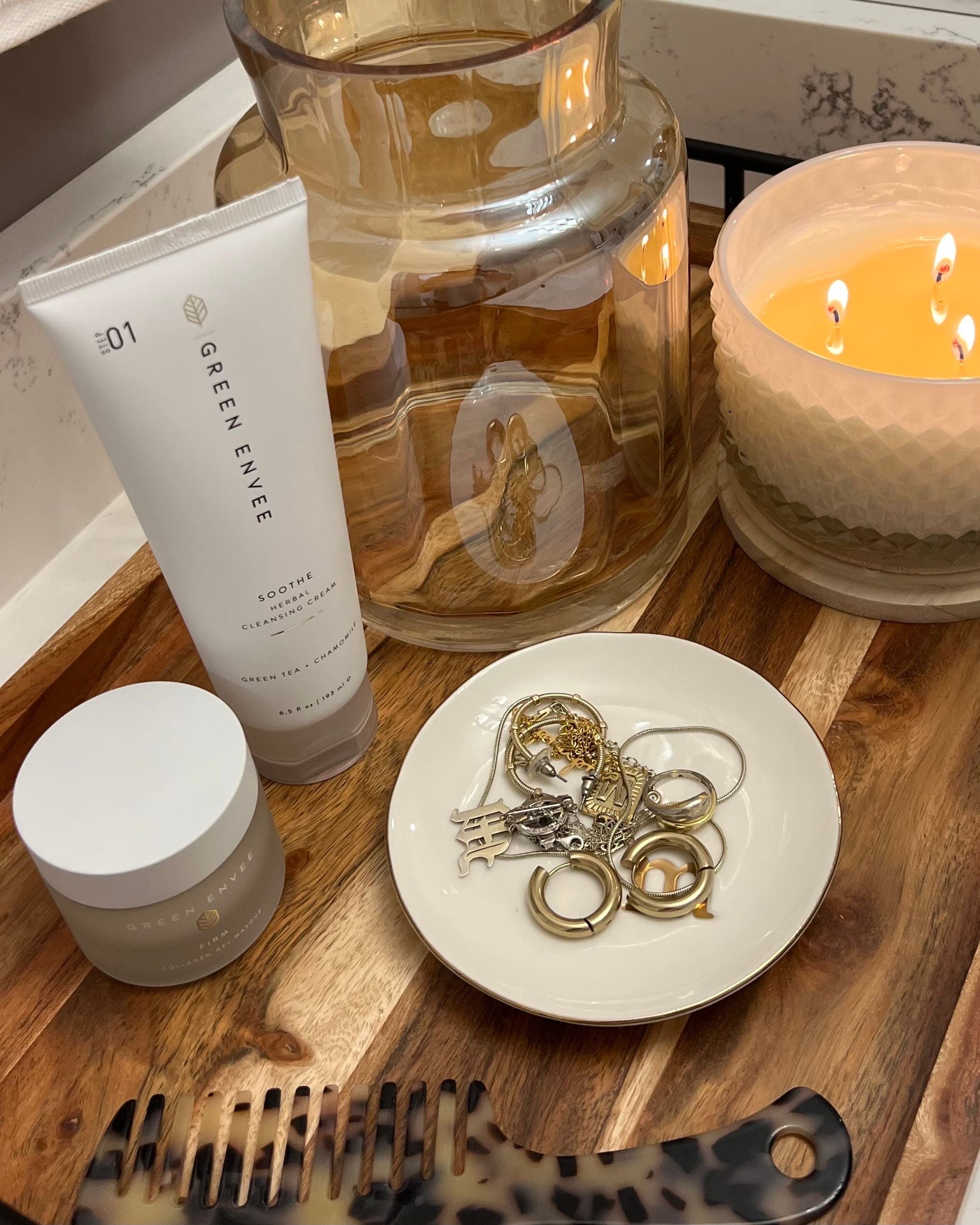 A styled tabletop arrangement featuring a white tube of Green Envee Body Butter, a small white jar, a white dish holding gold-toned jewelry, a large amber glass jar, and a lit candle on a wooden surface.