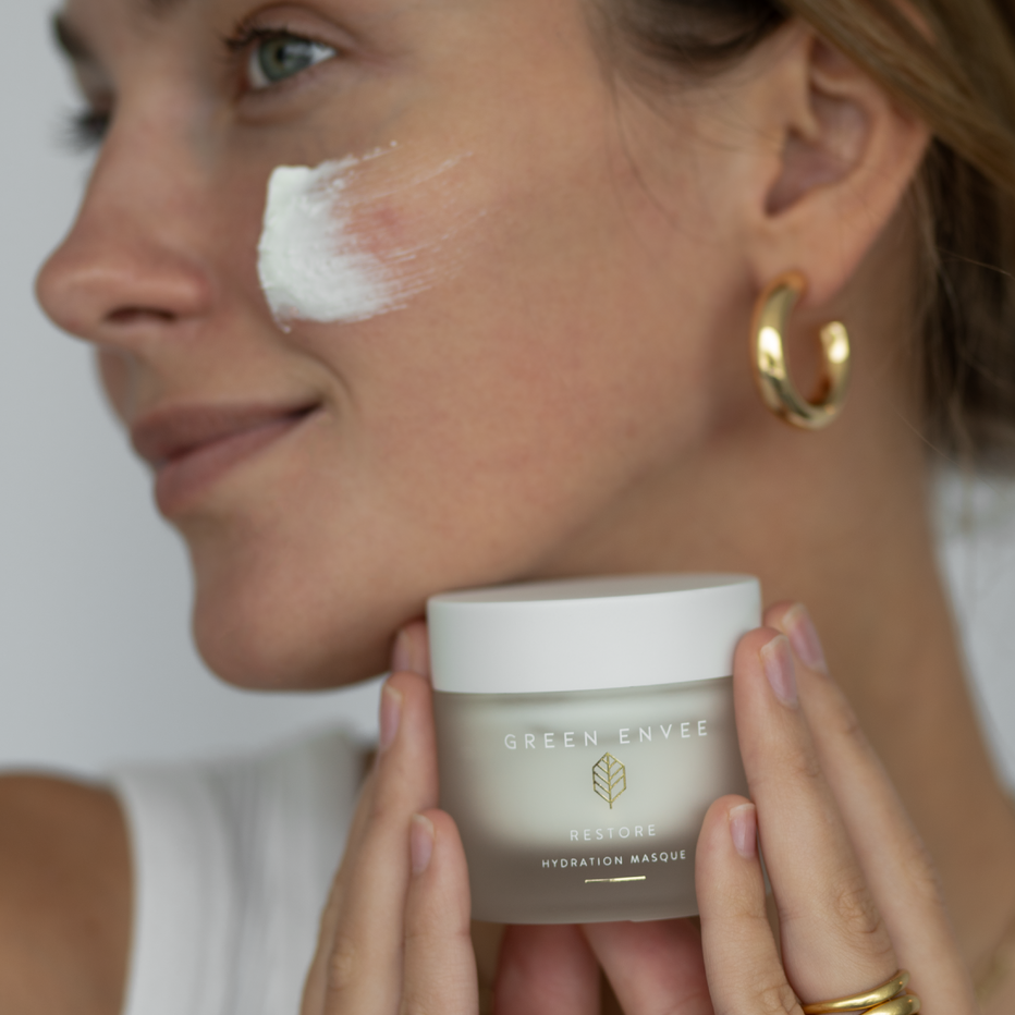 Woman holding a jar of 'Green Envee Restore Masque' product with a focus on her face.
