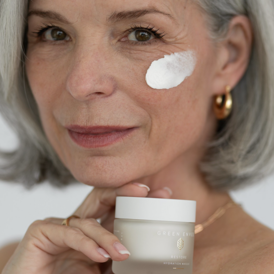 Woman applying cream to her face with a jar of the same cream in hand. Restore Masque